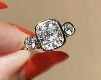 Puede incluir: Primer plano de un anillo de diamantes con una piedra central grande de talla cuadrada y dos piedras laterales redondas más pequeñas. El anillo está engastado en una banda de oro. Una joya.