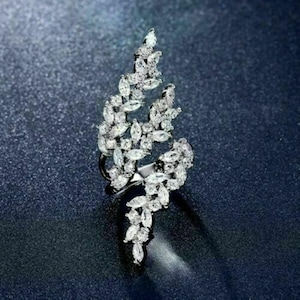 May include: A silver-toned ring with a leaf-like design, encrusted with numerous clear, sparkling gemstones. The ring features a curved, asymmetrical shape, with the gemstones arranged to mimic the form of leaves. The ring is set against a dark blue background.