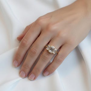 May include: A gold ring featuring three emerald-cut diamonds. The ring is set in a yellow gold band. The diamonds are clear and sparkling. The ring is on a hand with a white background.