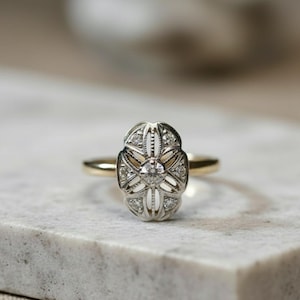 May include: A vintage-style ring with a gold band and an oval-shaped, silver-toned setting. The setting is adorned with small, sparkling gemstones and a central, larger stone. The ring is displayed on a light-colored, textured surface.