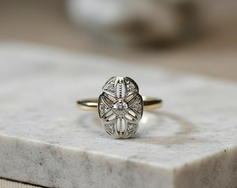 May include: A vintage-style ring with a gold band and an oval-shaped, silver-toned setting. The setting is adorned with small, sparkling gemstones and a central, larger stone. The ring is displayed on a light-colored, textured surface.