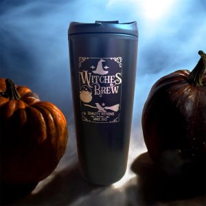 May include: A dark blue travel mug featuring a silver "Witches Brew" design, with a witch's hat, cauldron, and broom. Two pumpkins are on either side of the mug. The mug is designed for hot or cold drinks.