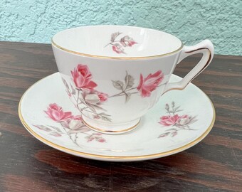 May include: White teacup and saucer set with a pink rose floral pattern and gold trim. The teacup has a curved handle and sits on a matching saucer. The set is likely vintage and suitable for serving tea.