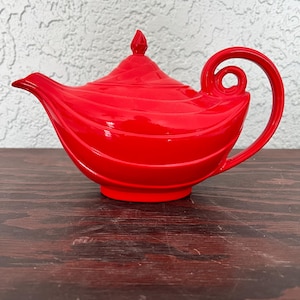Pode incluir: Uma chaleira vermelha vibrante com uma alça curva e uma tampa decorativa. A chaleira tem um acabamento brilhante e um design único em espiral. A chaleira está em uma superfície de madeira escura.