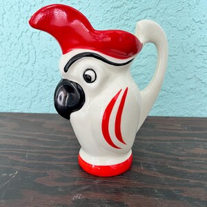Może przedstawiać: Biały ceramiczny dzbanek w kształcie papugi, z czerwonym czubem, czarnym dziobem i czerwonymi akcentami. Dzbanek ma zakrzywiony uchwyt i czerwoną podstawę. Ma około 15 cm wysokości.