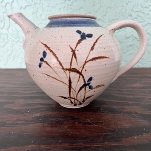Puede incluir: Una tetera de cerámica de color crema con un borde azul y un asa curva. La tetera está decorada con diseños florales marrones y azules. El pico está en el lado izquierdo.