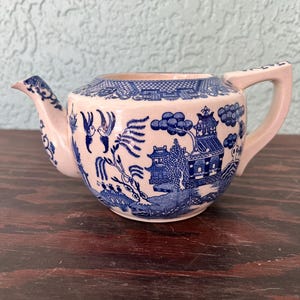 May include: A small, cream-colored teapot with a blue Willow pattern design. The teapot features a spout, handle, and lid, all adorned with the classic blue and white imagery of pagodas, bridges, and figures. The teapot is a vintage piece.