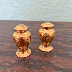 Puede incluir: Dos saleros y pimenteros antiguos de color dorado. Cada coctelera tiene un cuerpo bulboso, una pequeña base de pedestal y una tapa redondeada. Las cocteleras tienen una superficie texturizada y se exhiben sobre una superficie de madera oscura.