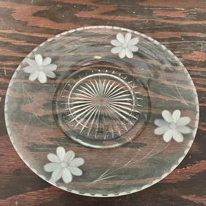 May include: A clear glass plate with a floral design. The plate features etched flowers and delicate leaf patterns around the rim. The center of the plate has a starburst design.