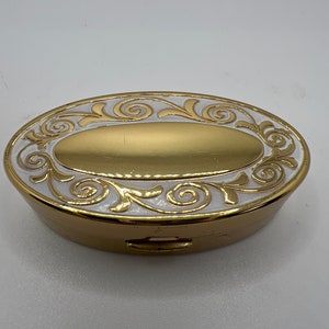 May include: A gold oval-shaped compact with a white enamel design featuring intricate swirls and floral motifs.
