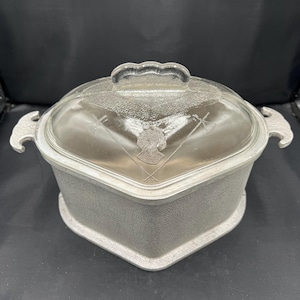 May include: A heart-shaped metal casserole dish with a textured silver-tone exterior and two side handles. The clear glass lid features a central embossed design of a profile silhouette and crossed axes, topped with a scalloped handle.