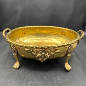 May include: A round brass bowl with a hammered interior finish and a rope-textured rim. The bowl features two side handles, three ornate feet shaped like animal paws, and decorative metal accents on the exterior sides of the vessel.