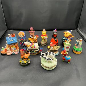 May include: A collection of decorative trinket boxes in various shapes and designs. The boxes feature colorful depictions of animals, characters, and scenes, including a rocking horse, a cow and calf, and a reindeer.