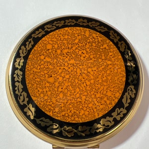 May include: A gold-toned compact mirror with a black and gold design. The mirror has a round, orange-brown surface with a speckled texture. The edge of the mirror is decorated with a black and gold leaf pattern.