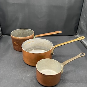 May include: Three copper saucepans with metallic interiors and long handles. The pans vary in size, featuring a warm copper exterior finish and a silver-toned interior lining. Each pan includes a single handle attached to the side of the round body.