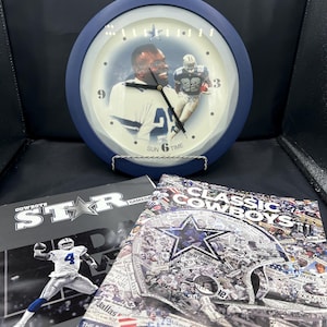 May include: A round wall clock with a dark blue frame featuring a Dallas Cowboys football player graphic, alongside two magazines. One magazine shows a player in a white jersey, and the other displays a collage of team photos and the text Classic Cowboys.