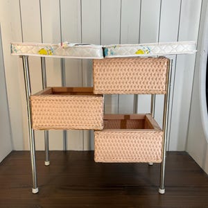 May include: A vintage baby changing table with a white and patterned changing pad. The table has three woven baskets in a light pink color and chrome legs. The changing pad is approximately 30 inches long.