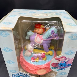 May include: A vintage Cabbage Patch Kids figurine set in its original box. The set features a girl riding a purple rocking horse with a blue mane and tail. A small white dog and colorful blocks are also included. The box has a light blue floral design.