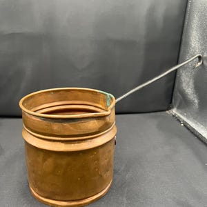 May include: A cylindrical copper pot with a smooth, warm metallic finish and a prominent horizontal ridge around the body. It features a long, thin, dark metal handle extending from the side and a small spout integrated into the top rim.