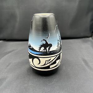 May include: A black and white ceramic vase with a Native American design. The vase features a landscape scene with a figure, mountains, and trees. The base has a geometric pattern.