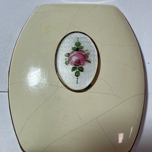 May include: A white oval compact with a gold rim and a painted pink rose with green leaves in the center. The compact has a cracked surface.