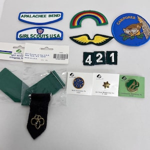 K&ouml;nnte beinhalten: Eine Sammlung von Girl Scout-Abzeichen, Anstecknadeln und Insignien. Enth&auml;lt Aufn&auml;her mit den Texten "APALACHEE BEND", "GIRL SCOUTS USA" und "CAMPOREE". Au&szlig;erdem ein Regenbogen-Aufn&auml;her, eine goldene Stern-Anstecknadel und eine gr&uuml;n-goldene Anstecknadel.