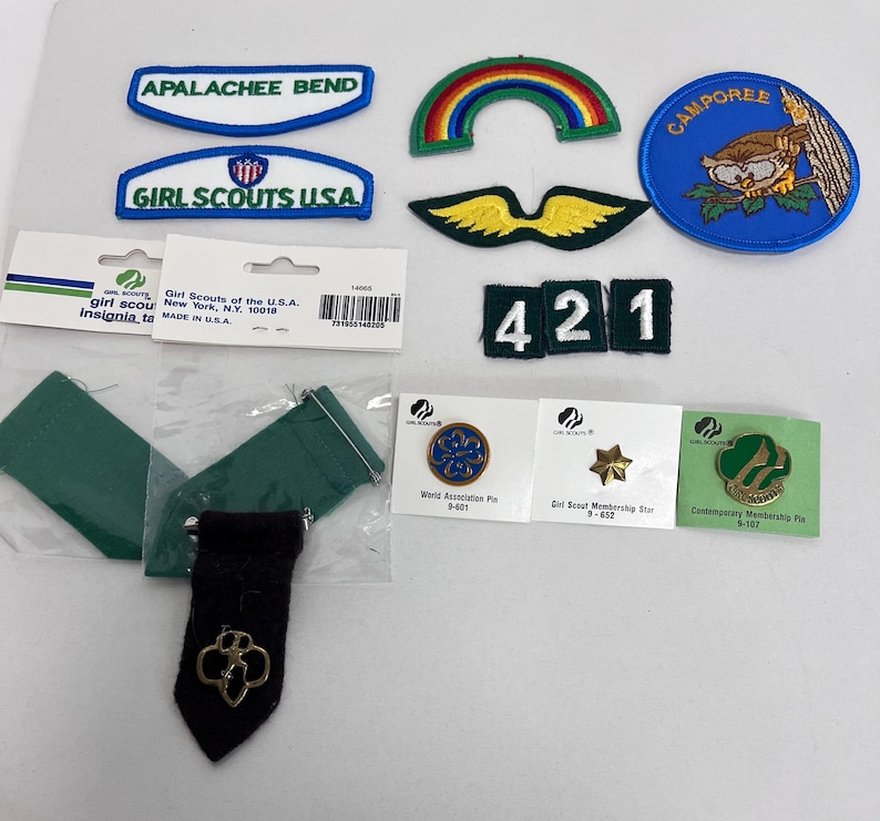 K&ouml;nnte beinhalten: Eine Sammlung von Girl Scout-Abzeichen, Anstecknadeln und Insignien. Enth&auml;lt Aufn&auml;her mit den Texten "APALACHEE BEND", "GIRL SCOUTS USA" und "CAMPOREE". Au&szlig;erdem ein Regenbogen-Aufn&auml;her, eine goldene Stern-Anstecknadel und eine gr&uuml;n-goldene Anstecknadel.