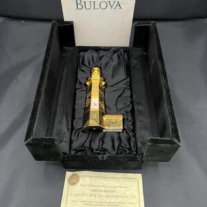 May include: A gold-toned Bulova miniature collectible, featuring a detailed clock tower design, presented in a black velvet-lined box. Includes a certificate of authenticity. The Bulova logo is visible on the box.