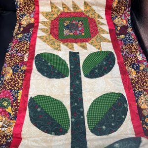 May include: A handmade quilt with a floral design. The quilt features a central flower motif with green leaves and a red border. The background is a cream color with a floral pattern.