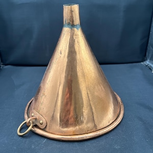 May include: A conical copper funnel with a polished metallic finish, featuring a wide circular base and a narrow top opening. A small brass ring is attached to the side of the base by a riveted metal tab, providing a handle for easy gripping.