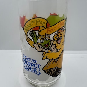 May include: A clear glass with a colorful image of Kermit the Frog and Fozzie Bear from The Great Muppet Caper. The image shows Kermit holding a microphone and Fozzie Bear wearing a hat with the word "Press" on it.