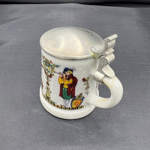 May include: White ceramic beer stein with a hinged metal lid and handle. The stein features a colorful illustration of a person playing a musical instrument. The design includes floral and decorative elements. The stein is approximately 4 inches tall.