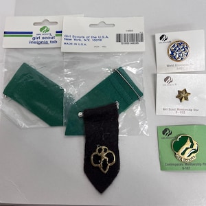 K&ouml;nnte beinhalten: Verschiedene Girl Scout-Insignien und -Anstecknadeln. Enth&auml;lt ein schwarzes Insignien-Tab mit goldenem Emblem, gr&uuml;ne Filztaschen und verschiedene Anstecknadeln mit dem Girl Scout-Logo und einem Stern. Die Artikel befinden sich auf wei&szlig;en Karten und in Kunststoffverpackungen.