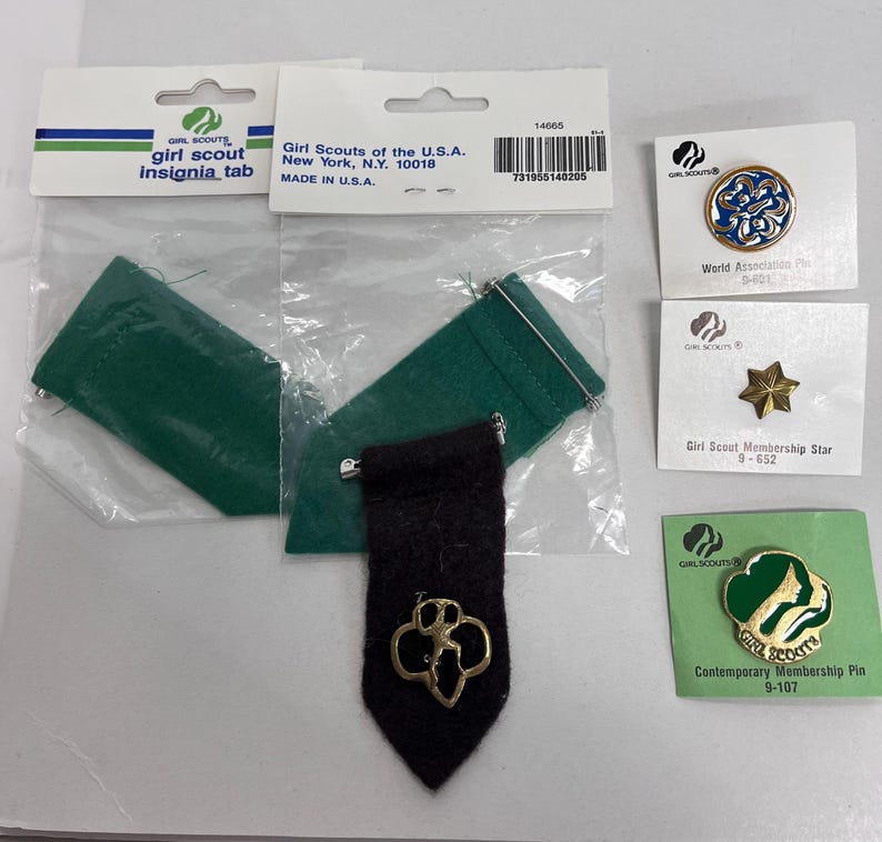 K&ouml;nnte beinhalten: Verschiedene Girl Scout-Insignien und -Anstecknadeln. Enth&auml;lt ein schwarzes Insignien-Tab mit goldenem Emblem, gr&uuml;ne Filztaschen und verschiedene Anstecknadeln mit dem Girl Scout-Logo und einem Stern. Die Artikel befinden sich auf wei&szlig;en Karten und in Kunststoffverpackungen.