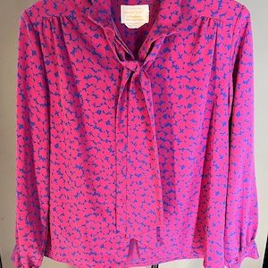 May include: A long-sleeved blouse in a vibrant pink hue, adorned with a repeating pattern of blue shapes. The blouse features a tie-neck design and a label that reads "Country Sophisticates". The sleeves have buttoned cuffs.