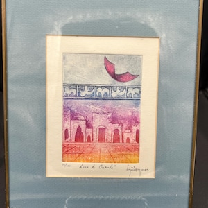 May include: A framed art print titled "Luna de Oriente" with a colorful cityscape scene. The print features a red and purple sky, a row of buildings, and a pink crescent shape. The frame is gold with a light blue mat.