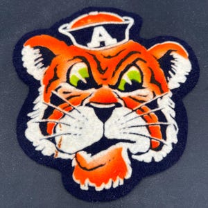 May include: A felt patch featuring the head of an orange and white tiger with black stripes and green eyes. The tiger is wearing an orange cap with a white letter A on the front. The entire design is outlined in a dark navy blue felt border.