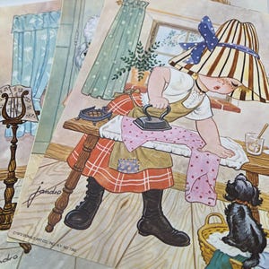 May include: A stack of vintage prints featuring a whimsical illustration of a person ironing clothes. The person wears a striped hat and apron, with a dog nearby. The prints have a warm color palette and include text from the artist.
