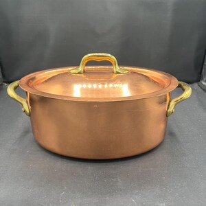 May include: A copper-colored pot with a matching lid. The pot has two side handles and a top handle, all in a gold tone. The pot is round and sits on a dark surface. This cookware is suitable for cooking.