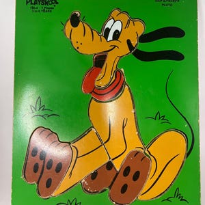May include: A vintage Playskool wooden puzzle featuring Pluto, a cartoon dog from Disney's animated films. The puzzle has 7 pieces and is recommended for children ages 3 to 5 years old. The puzzle is set against a green background with grass and features Pluto sitting with his tongue sticking out.