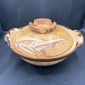 May include: A round ceramic donabe pot with a matching lid, featuring a mottled golden-brown glaze and a hand-painted bamboo leaf design. The pot includes two side handles and a central knob on the lid, showcasing a textured, earthy finish.