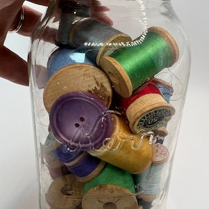 May include: A clear glass jar filled with colorful spools of thread and a purple button. The spools are various colors, including green, blue, and red. The jar has a metal lid and is held by a hand.