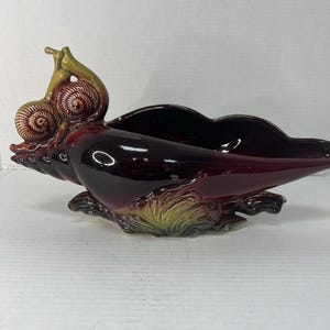 May include: A burgundy ceramic seashell planter with two snails on top. The planter has a green and yellow glaze and is shaped like a seashell.