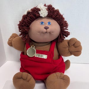 May include: A Cabbage Patch Kids plush doll with brown fur and a red jumpsuit. The doll has blue eyes, a brown face, and a white and brown hairstyle. A gold-colored medallion hangs from its neck.