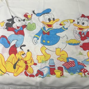 May include: A white cotton fabric with a colorful cartoon print of Mickey Mouse, Donald Duck, Daisy Duck, and Pluto having a picnic. The characters are surrounded by picnic items like a basket, a thermos, and a plate of pie.