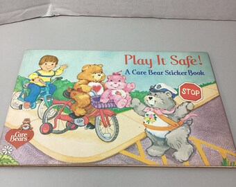 Puede incluir: Un libro de pegatinas de los Osos Amorosos titulado "Play It Safe!" con un niño montando en bicicleta, tres Osos Amorosos montando en triciclo y un Oso Amoroso con uniforme de policía de pie cerca de una señal de stop.
