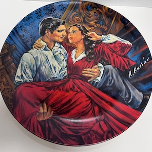 May include: A round decorative plate featuring a colorful illustration of a man in a white shirt and a person in a long red dress. The artwork depicts the two figures in an embrace against a dark blue and gold background, signed R. Kursár.
