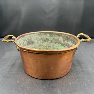 May include: A small, antique copper bowl with two brass handles. The bowl has a rounded shape and a slightly tarnished, aged appearance. The interior shows green oxidation, adding to its vintage charm. The bowl is likely used for decorative purposes.