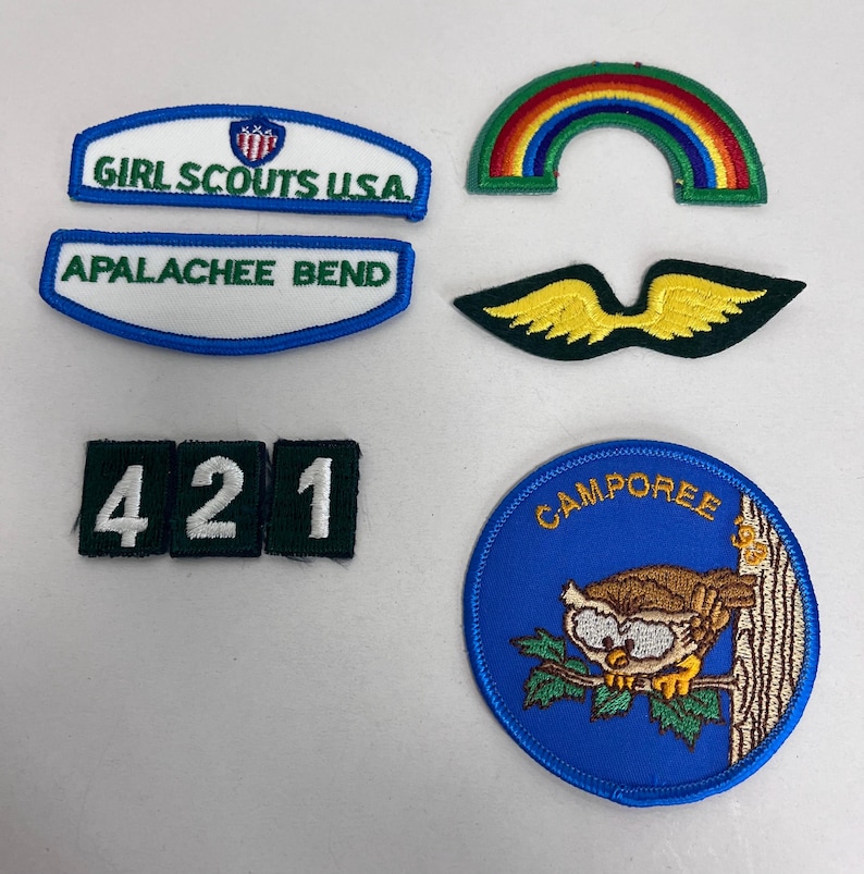K&ouml;nnte beinhalten: Eine Sammlung gestickter Aufn&auml;her, darunter ein Regenbogen, Fl&uuml;gel und ein blauer Kreis mit einer Eule. Text enth&auml;lt "GIRL SCOUTS USA", "APALACHEE BEND", "421" und "CAMPOREE". Die Aufn&auml;her sind in verschiedenen Formen und Farben.