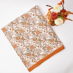 May include: A white cotton throw blanket with an orange floral pattern and a burnt orange border. The blanket is folded and laid flat on a white surface.